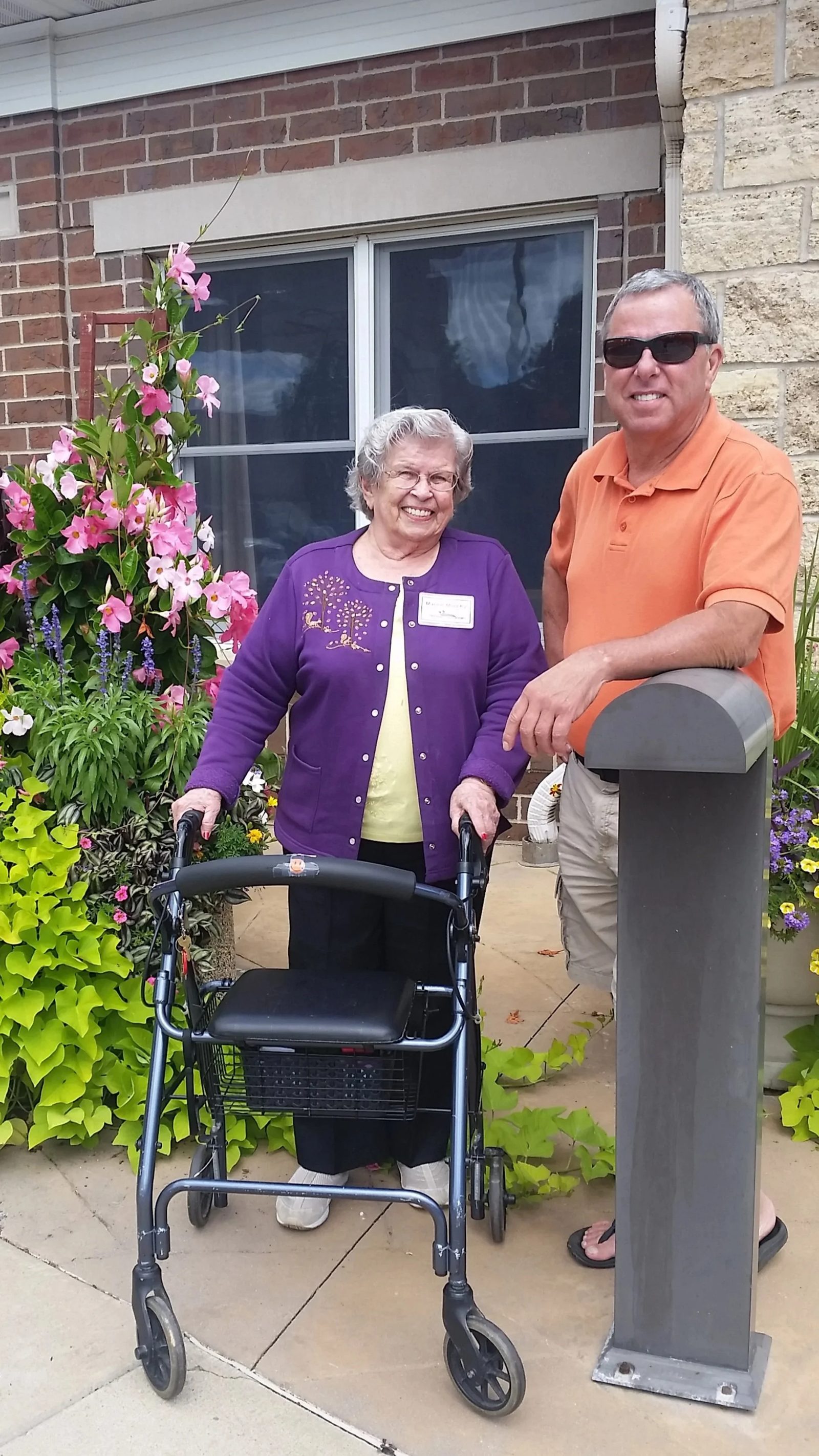 Kevin Murphy smiling beside an older woman with her walker outside a brick building with flowers
