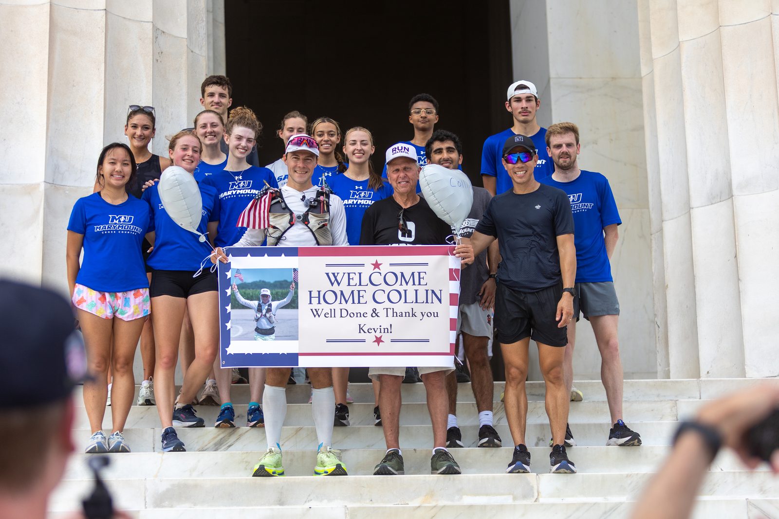Runners and supporters on the Lincoln Memorial steps holding a banner that reads Welcome Home Collin, Well Done and Thank You Kevin