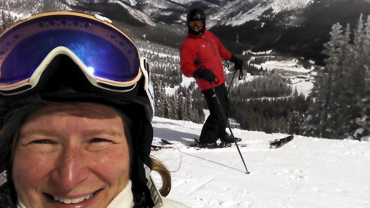 A smiling skier in goggles with another skier on the slope behind